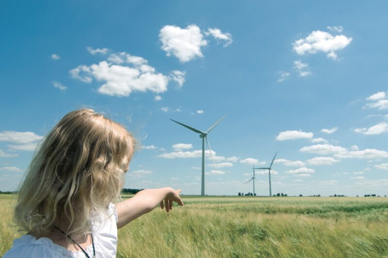 Eine junge Frau zeigt Windkraftanlagen auf einer grünen Wiese vor blauem Himmel als Zeichen für Bürgerenergie.