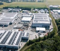 Luftaufnahme eines Industriegeländes mit großflächigen Photovoltaikanlagen auf mehreren Hallendächern. Zwischen den Gebäuden liegen Zufahrten und Lagerflächen, am Rand Bäume und Felder.