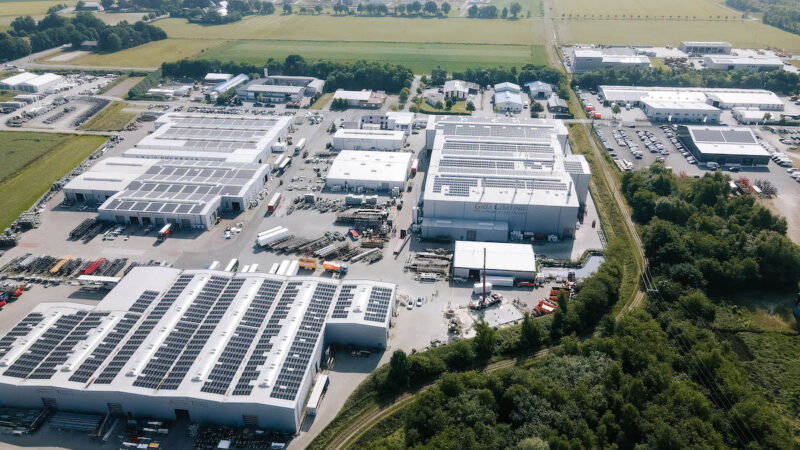 Luftaufnahme eines Industriegeländes mit großflächigen Photovoltaikanlagen auf mehreren Hallendächern. Zwischen den Gebäuden liegen Zufahrten und Lagerflächen, am Rand Bäume und Felder.