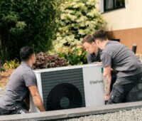 Das Bild zeigt drei grau gekleidete Männer in Arbeitskleidung, die gemeinsam zu einem Haus eine Wärmepumpe tragen.