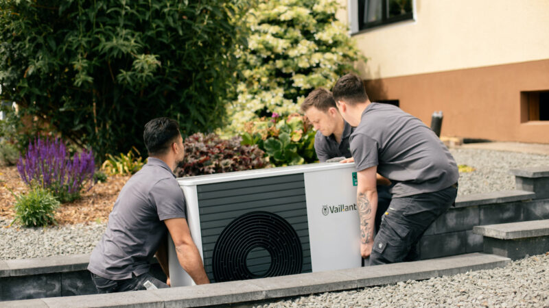 Das Bild zeigt drei grau gekleidete Männer in Arbeitskleidung, die gemeinsam zu einem Haus eine Wärmepumpe tragen.