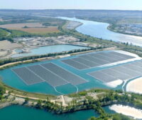 Die Luftaufnahme zeigt Reihen von Solarmodulen auf einem von mehreren Seen. Der See ist durch einen Erdwall und eine Straße vom Fluss Rhone getrennt