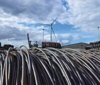 Stahl-Seile im Vordergrund, hinten ein Industriegelände mit einer Windkraftanlage. Erneuerbare Energien für grünen Stahl.
