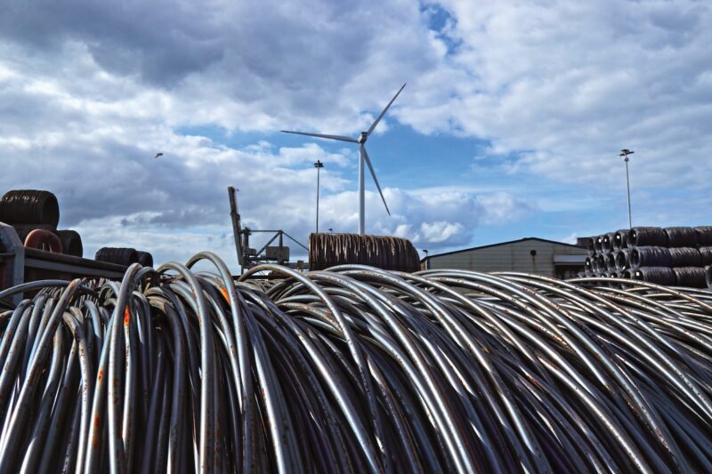 Stahl-Seile im Vordergrund, hinten ein Industriegelände mit einer Windkraftanlage. Erneuerbare Energien für grünen Stahl.