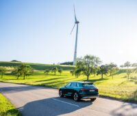 Zusehen ist ein Audi e-tron Elekotrauto bei einer Testfahrt mit einem Windrad im Hintergrund. Audi testet zurzeit das Batterierecycling.