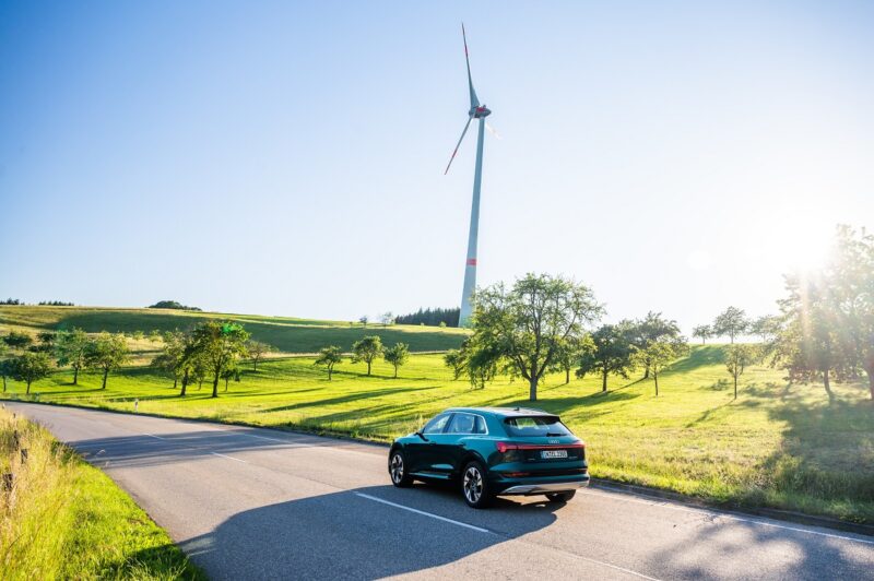 Zusehen ist ein Audi e-tron Elekotrauto bei einer Testfahrt mit einem Windrad im Hintergrund. Audi testet zurzeit das Batterierecycling.