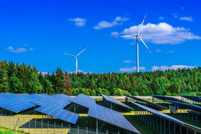 Ein Solarpark neben einem Wald und Windenergieanlagen.