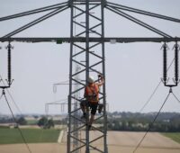 Zu sehen ist ein Arbeiter, der auf einen Strommast einer Hochspannungsleitung klettert. Die Bundesnetzagentur setzt die Eigenkapitalkosten für Strom- und Gasnetzbetreiber fest.