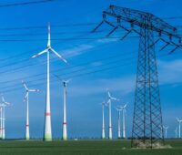 Im Bild Windenergieanlagen und ein Strommast, Auktionen sollen die Abregelung von Grünstrom vermindern.