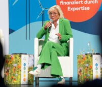 LEE-Vorsitzende Bärbel Heidebroek sitzt im grünen Hosenanzug auf einem Podium und spricht in ein Handmikrofon. Im Hintergrund sind Windenergie-Motive zu sehen.