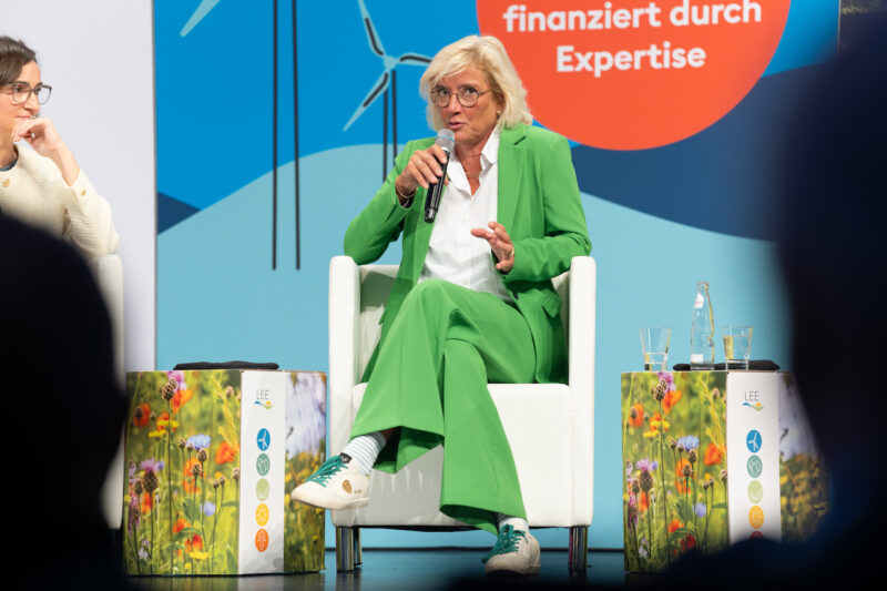 LEE-Vorsitzende Bärbel Heidebroek sitzt im grünen Hosenanzug auf einem Podium und spricht in ein Handmikrofon. Im Hintergrund sind Windenergie-Motive zu sehen.