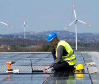 Zu sehen ist die Montage einer Photovoltaik-Dachanlage, im Hintergrund sind Windenergieanlagen. Die BMWi-Langfristszenarien unterschätzen laut BEE die nationalen Potenziale für erneuerbare Energien.