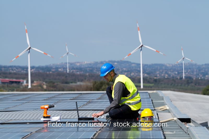 Zu sehen ist die Montage einer Photovoltaik-Anlage vor Windkraftanlagen, deren Ausbau von der beihilferechtlichen Genehmigung des EEG 2021 abhängt.