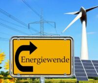 Ein gelbes Ortseingangsschild mit der schwarzen Aufschrift Energiewende im Linksbogen eines schwarzen Pfeils nach oben, im Hintergrund Strommasten, eine Windenergieanlage und rechts PV-Module sowie links Sonnenblumen.