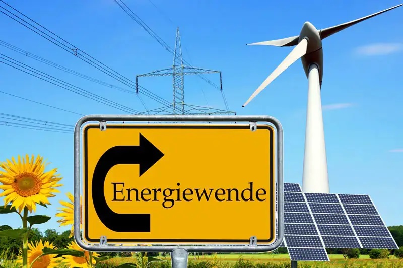 Ein gelbes Ortseingangsschild mit der schwarzen Aufschrift Energiewende im Linksbogen eines schwarzen Pfeils nach oben, im Hintergrund Strommasten, eine Windenergieanlage und rechts PV-Module sowie links Sonnenblumen.