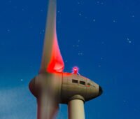 Nachtaufnahme (Langzeitbelichtung) der Gondel einer Enercon-Windenergieanlage Typ E-101 Mit roter Nachtbeleuchtung vor Sternenhimmel.