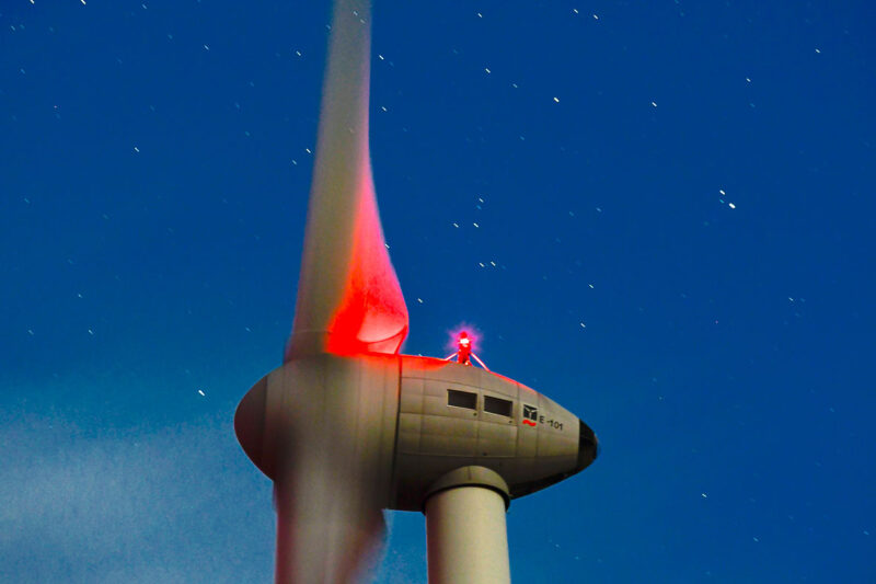 Nachtaufnahme (Langzeitbelichtung) der Gondel einer Enercon-Windenergieanlage Typ E-101 Mit roter Nachtbeleuchtung vor Sternenhimmel.