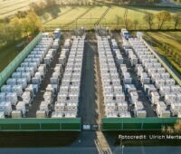 Luftaufnahme des Batteriespeichers Bollingstedt mit zahlreichen weißen Speichercontainern in parallelen Reihen, eine grüne Schutzwand umschließt das Betriebsgelände, umgeben von Feldern und Bäumen.
