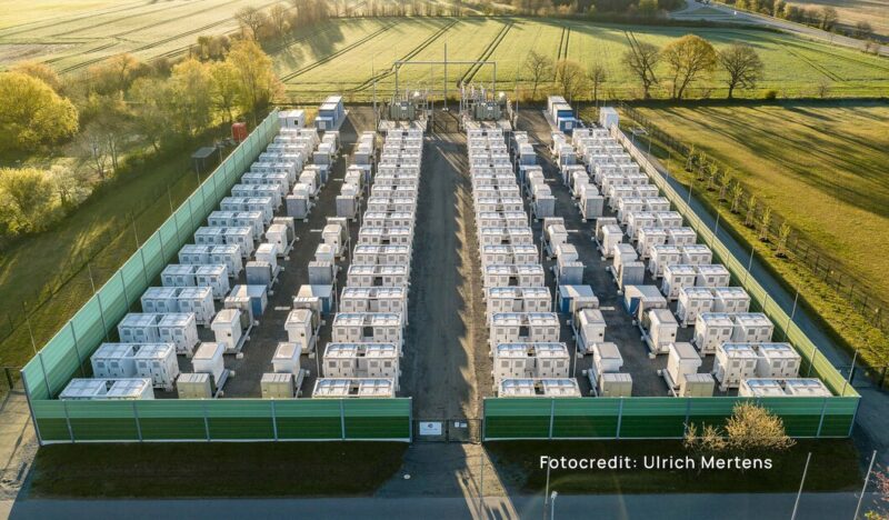 Luftaufnahme des Batteriespeichers Bollingstedt mit zahlreichen weißen Speichercontainern in parallelen Reihen, eine grüne Schutzwand umschließt das Betriebsgelände, umgeben von Feldern und Bäumen.
