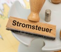 Nahaufnahme eines Holzstempels mit der schwarzen Aufschrift Stromsteuer liegt auf Unterlagen auf einem Tisch.