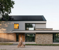 Modernes zweigeschossiges Einfamilienhaus mit einer Photovoltaikanlage auf dem geneigten Dach. Vor dem Gebäude läuft eine erwachsene Person mit einem Kleinkind vorbei. Links steht ein großer Baum, im Hintergrund ist eine Wohnstraße zu sehen.