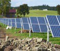 Auf einer hügeligen grünen Wiese stehen Reihen von aufgeständerten Solarmodulen vor einem Stall mit rotem Dach