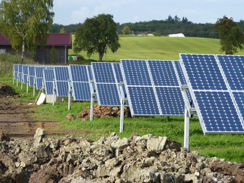 Auf einer hügeligen grünen Wiese stehen Reihen von aufgeständerten Solarmodulen vor einem Stall mit rotem Dach