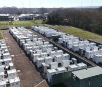 Das Foto zeigt in drei Reihen hellgraue Batteriespeichersysteme hintereinander aus der Luft, rechts und links der Anlage sind Baumreihen, im Hintergrund Gebäude und ein grauer Himmel.