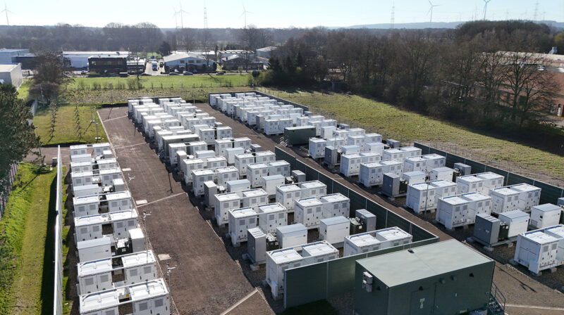 Das Foto zeigt in drei Reihen hellgraue Batteriespeichersysteme hintereinander aus der Luft, rechts und links der Anlage sind Baumreihen, im Hintergrund Gebäude und ein grauer Himmel.