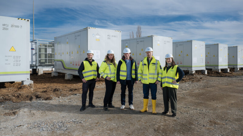 Das Bild zeigt fünf Personen mit weißen Schutzhelmen und neongelben Jacken und Westen vor mehreren Batteriespeicher-Systemen, schräg dahinter.