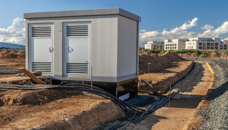 Graue Netzstation auf einer Baustelle, aus deren Fundament mehrere dicke Erdkabel herausgeführt und in einen offenen Graben verlegt werden. Im Hintergrund Erdaushub und mehrstöckige Gebäude.