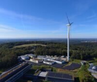 Luftaufnahme zeigt Flachgebäude mit PV-Dächern, Freiflächen-Solar und Windenergieanlage