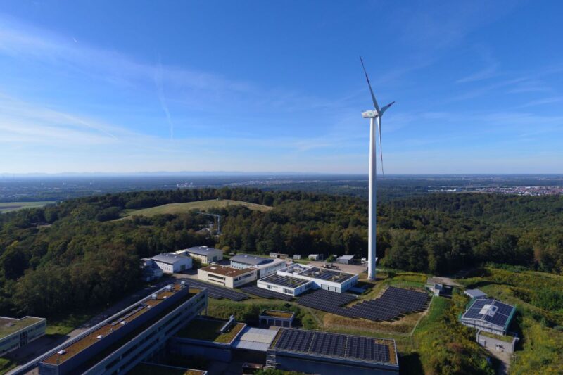 Luftaufnahme zeigt Flachgebäude mit PV-Dächern, Freiflächen-Solar und Windenergieanlage