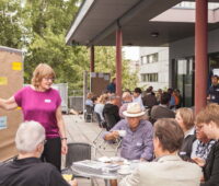 Menschen sitzen an Tischen vor einem Gebäude und trinken Kaffee. Trainer stehen an Schautafeln und erläutern