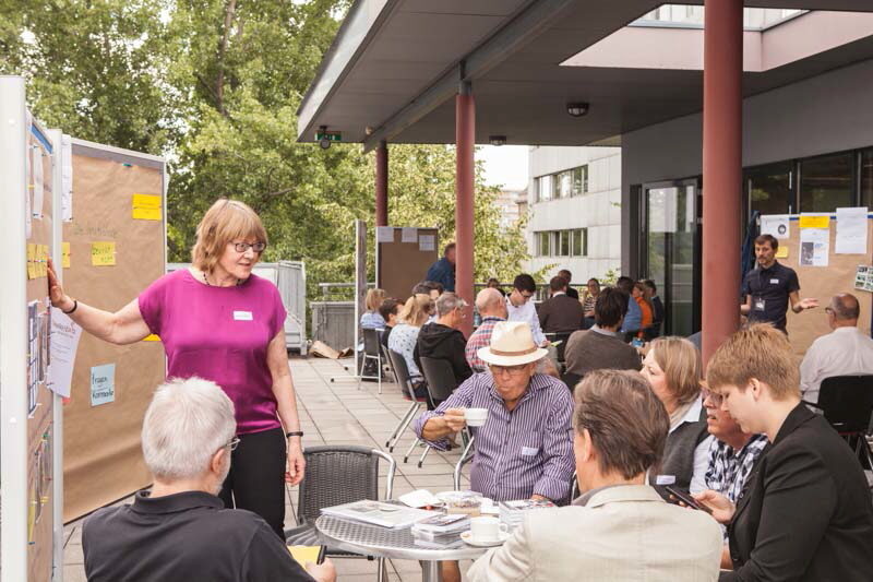 Menschen sitzen an Tischen vor einem Gebäude und trinken Kaffee. Trainer stehen an Schautafeln und erläutern