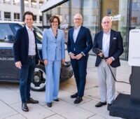 Das Foto zeigt vier Personen in Anzügen in Blautönen stehend vor einem dunkelblauen VW Bulli ID. Buzz mit weißem Dach, daneben eine Ladesäule. Die rechte Person hält ein Ladekabel in der Hand. Im Hintergrund Gebäudefassaden.