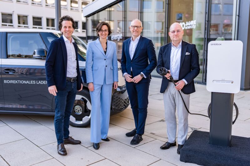 Das Foto zeigt vier Personen in Anzügen in Blautönen stehend vor einem dunkelblauen VW Bulli ID. Buzz mit weißem Dach, daneben eine Ladesäule. Die rechte Person hält ein Ladekabel in der Hand. Im Hintergrund Gebäudefassaden.