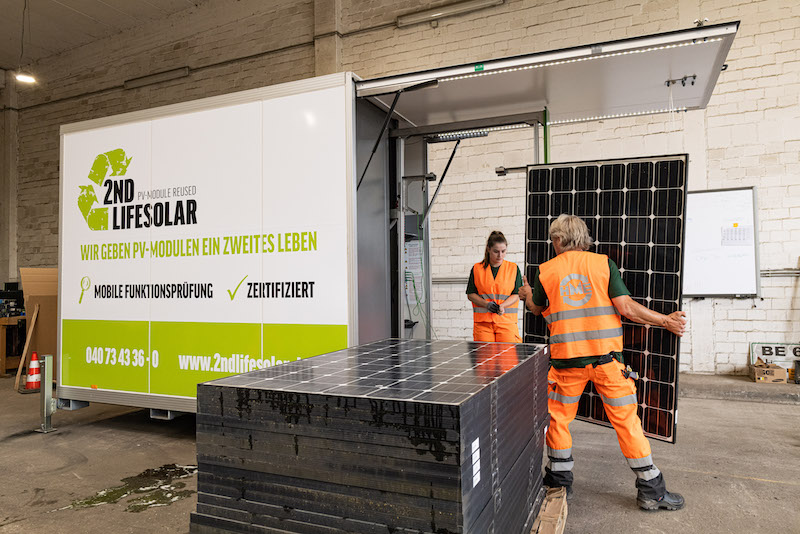 ein grün-weißer Container mit geöffneter Klappe, ein Stapel Photovoltaik-Module und zwei Menschen mit orangefarbenen Anzügen: der mobile Teststand des Recycling-Anbieters Buhck