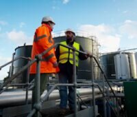Zwei Männer auf einer Industriebrücke vor großen Behältern einer Biogas-Anlage