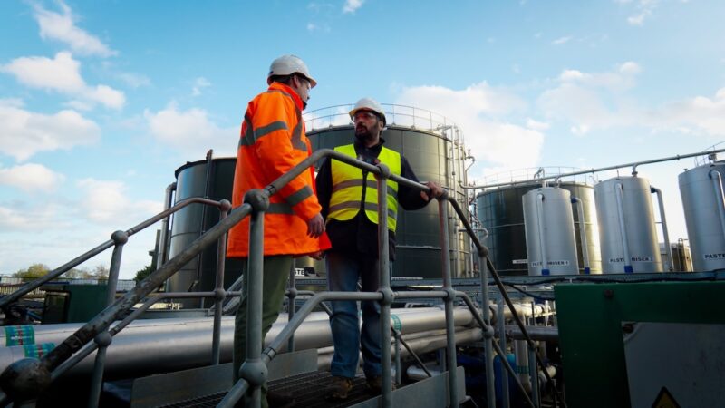 Zwei Männer auf einer Industriebrücke vor großen Behältern einer Biogas-Anlage