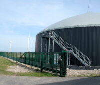 Ein Biogasfermenter steht im Vordergrund, daneben Windkraftanlagen unter blassblauem Himmel.