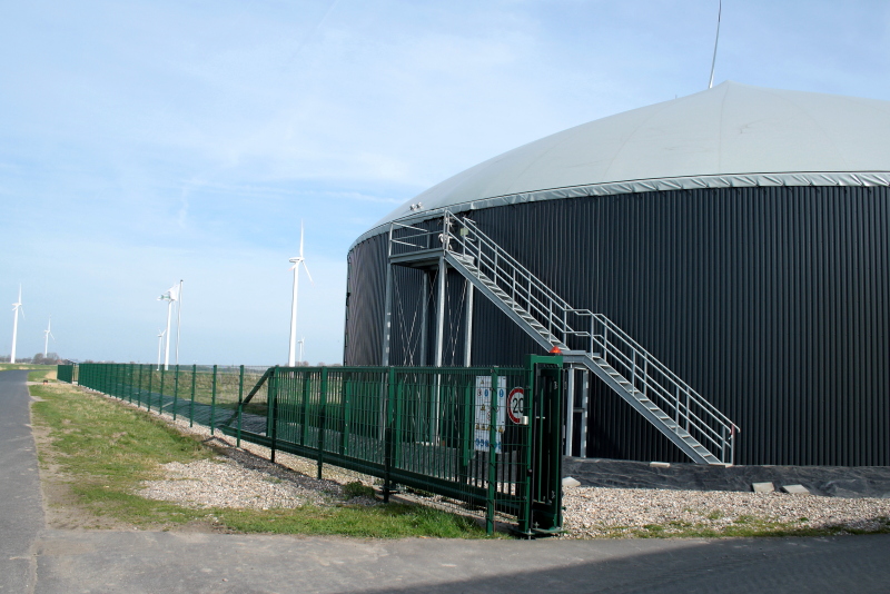 Ein Biogasfermenter steht im Vordergrund, daneben Windkraftanlagen unter blassblauem Himmel.