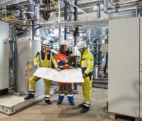 Blick in die Pyrolyse-Anlage bei RWE: Zwei Männer mit gelben Schutzanzügen und eine Frau mit orangefarbenem Schutzanzug stehen zwischen grauen Gehäusen und halten einen Plan in der Hand.