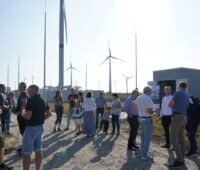 Menschen bei einer Einweihung einer Wärmezentrale unter blauem Himmel und Windenergieanlagen im Hintergrund