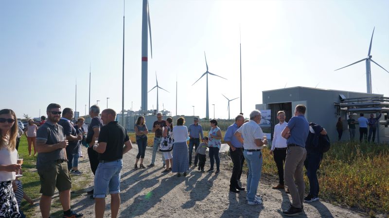 Menschen bei einer Einweihung einer Wärmezentrale unter blauem Himmel und Windenergieanlagen im Hintergrund