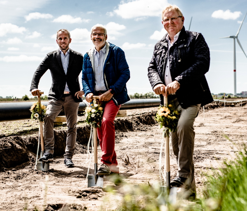 Drei Herren beim Spatenstich in Nordfriesland. Im Hintergrund Windkraftanlagen.