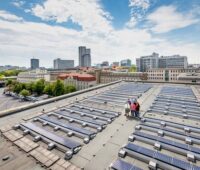 Bürger stehen zwischen Solarmodulen auf einem Flachdach in einer Stadt.