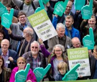 Gruppe von Menschen aus der Vogelperspektive demonstriert mit Pappschildern und überdimensionalen grünen Schaumstoffhänden für Bürgerenergie wie Energy Sharing.