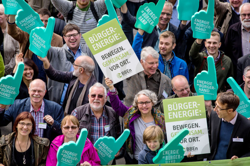 Gruppe von Menschen aus der Vogelperspektive demonstriert mit Pappschildern und überdimensionalen Schaumstoffhänden für Bürgerenergie, u.a. Energy Sharing.