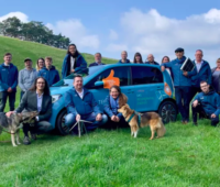 Auf einer grünen Wiese stehen die Mitarbeiter:innen der Teckwerke Bürgerenergie eG um ein blaues Auto in einer Hügellandschaft.